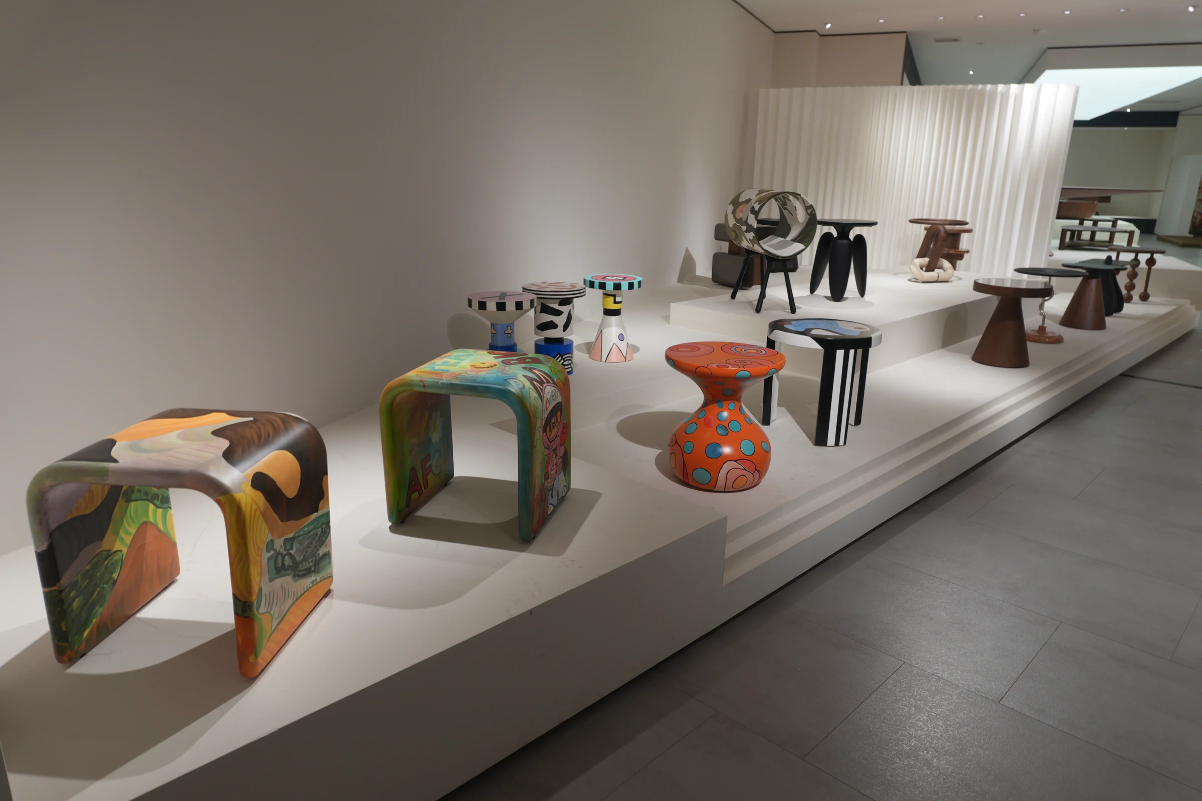 Colourful painted stools and sculptural side tables arranged on white showroom platforms